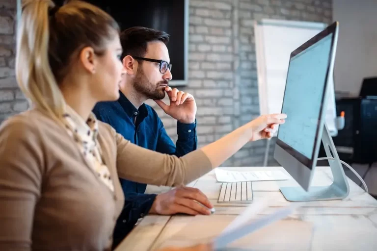 A woman points to the screen of a computer that a man works at in a modern office setting.