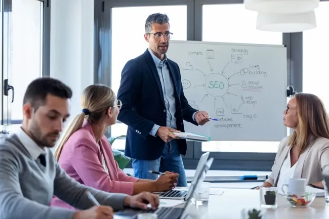 handsome-businessman-explaining-a-project-to-his-c-2024-09-12-17-41-19-utc-min A businessman explains an SEO project to his team on a white board in a meeting space.
