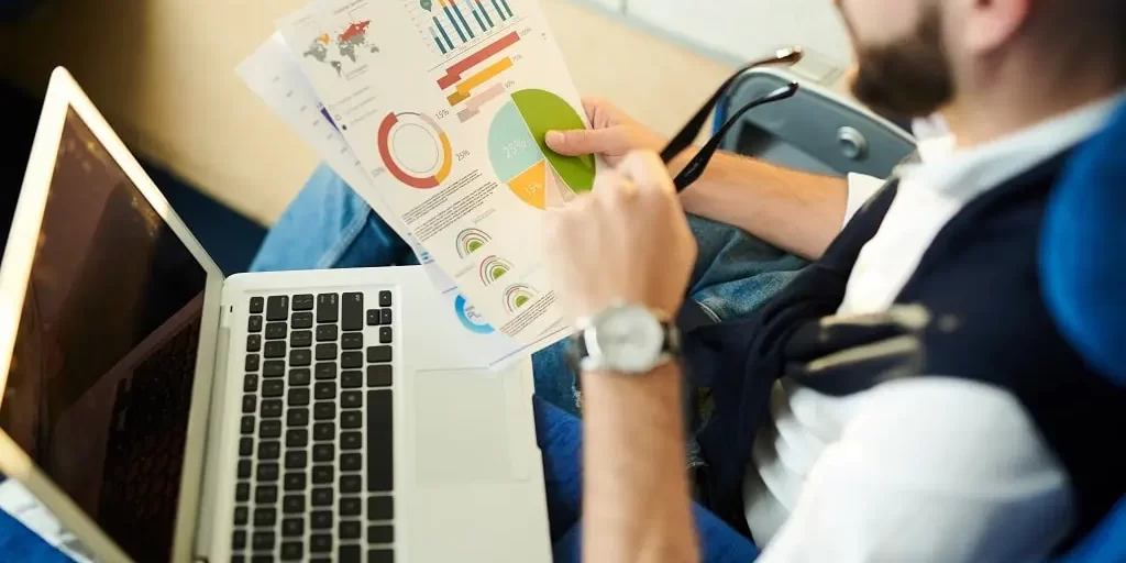 A businessman in an airplane seat studies a graph of data he holds in one hand, with his glasses in the other. His laptop sits on his lap.